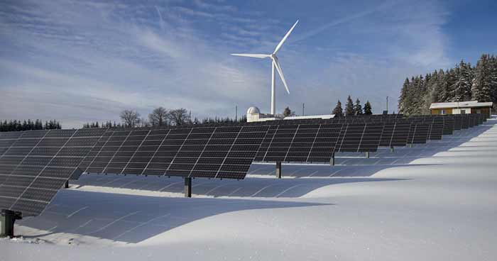 Solpaneler i snötäckt landskap med vindkraftverk i bakgrunden